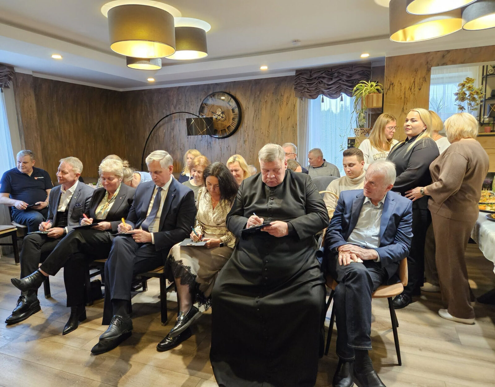Žmonės sėdi eilėmis šviesioje salėje ir klausosi susitikimo.